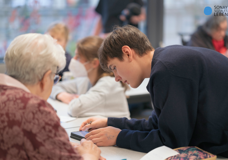 Ein Jugendlicher zeigt einer älteren Dame etwas an einem Smartphone. Im Hintergrund sind weitere Menschen verschiedenen Alters zu sehen.
